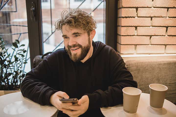 En leende man med skägg sitter vid ett runt bord framför ett fönster med kaffemugg och telefon.